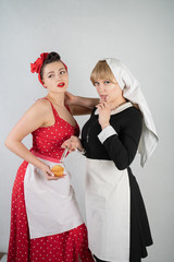 two cute girlfriends dressed up for a costume cosplay party. one girl is dressed as a housewife in pinup style, the second woman is dressed as a nun with a syringe. they stand on a white background.