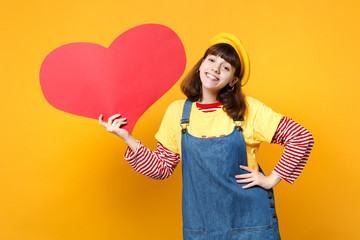 Pretty funny girl teenager in french beret and denim sundress holding big empty blank red heart isolated on yellow background in studio. People sincere emotions, lifestyle concept. Mock up copy space.