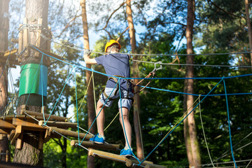 Sporty, young, cute boy in white t shirt spends his time in adventure rope park in helmet and safe equipment in the park in the summer. Active lifestyle concept, summer camp