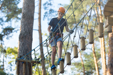 Sporty, young, cute boy in white t shirt spends his time in adventure rope park in helmet and safe equipment in the park in the summer. Active lifestyle concept, summer camp