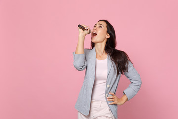 Portrait of attractive young woman in striped jacket looking up, sing song in microphone isolated on pink pastel background in studio. People sincere emotions, lifestyle concept. Mock up copy space.