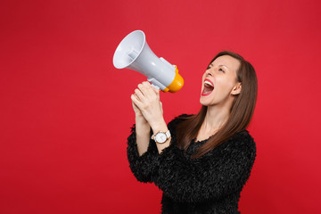 Portrait of joyful young girl in black fur sweater looking up, screaming on megaphone isolated on bright red wall background in studio. People sincere emotions, lifestyle concept. Mock up copy space.