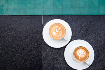Two white cups of tasty cappucino with art latte on the edge of swimming pool.