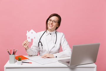 Female doctor sit at desk work on computer with medical document hold house in hospital isolated on pastel pink wall background. Woman in medical gown glasses stethoscope. Healthcare medicine concept.