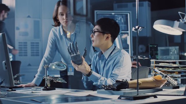 Technological Prosthetic Robot Arm is Tested by Two Professional Development Engineers in a High Tech Research Laboratory with Modern Futuristic Equipment. Man and Female Compare Data on a PC