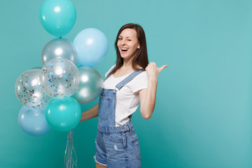 Fototapeta premium Cheerful young woman in denim clothes blinking, pointing thumb aside celebrating hold colorful air balloons isolated on blue turquoise wall background. Birthday holiday party, people emotions concept.