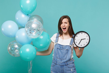Nervous screaming young woman in denim clothes holding round clock while celebrating with colorful air balloons isolated on blue turquoise background. Birthday holiday party, people emotions concept.