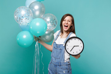 Funny screaming young woman in denim clothes holding round clock and celebrating with colorful air...