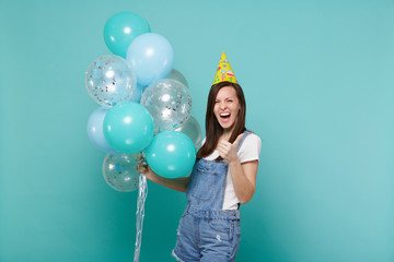 Screaming young woman in denim clothes, birthday hat showing thumb up celebrating, holding colorful air balloons isolated on blue turquoise background. Birthday holiday party, people emotions concept.