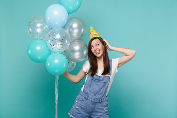 Funny woman in birthday hat showing tongue, looking up, put hand on head celebrating, hold colorful air balloons isolated on blue turquoise background. Birthday holiday party, people emotions concept.