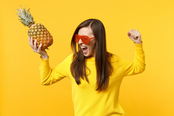 Screaming crazy young woman in funny glasses clenching fist holding fresh ripe pineapple fruit isolated on yellow orange background. People vivid lifestyle, relax vacation concept. Mock up copy space.