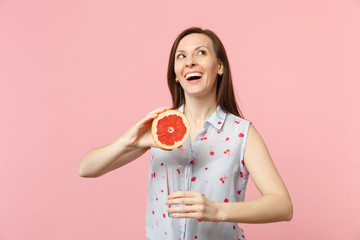 Dreamful young woman in summer clothes looking up holding half of fresh ripe grapefruit glass cup isolated on pink pastel background. People vivid lifestyle relax vacation concept. Mock up copy space.