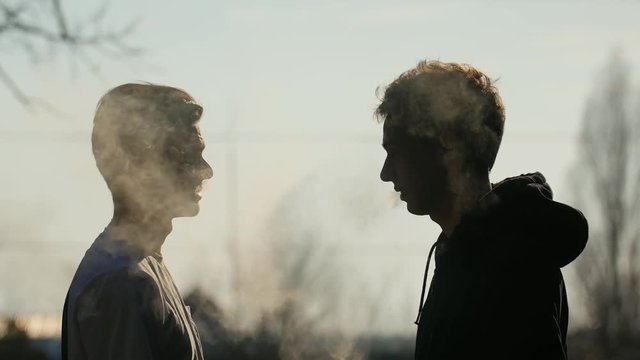 Silhouette of two talkative friends that smoking and vaping outdoors
