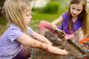 Two funny little girls playing in a large wet mud puddle on sunny summer day. Children getting dirty while digging in muddy soil.