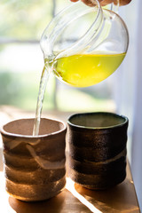 Closeup of person pouring green tea in cups in summer in home breakfast morning with yellow Sencha color and stream by window two Japanese teacups