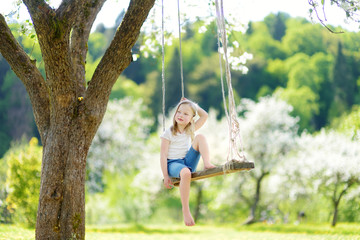 Cute little girl having fun on a swing in blossoming old apple tree garden outdoors on sunny spring day.