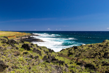 Hawaiian coastline near South Point on the Big Island. Volcanic rocks and grass on the hill in foreground; Pacific ocean in the distance. 