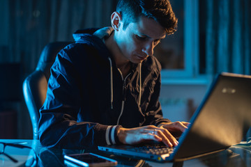 Fototapeta premium Young man worker working on a laptop at night. Freelancer designer or system administrator stayed late at work.
