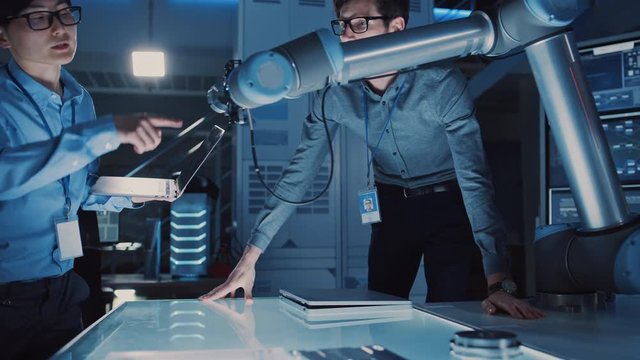 Diverse Team of Engineers with Laptop and a Tablet Analyse and Discuss How a Futuristic Robotic Arm Works and Moves a Metal Object. They are in a High Tech Research Laboratory with Modern Equipment.