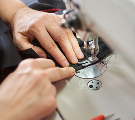 Obraz na płótnie Canvas Top view of woman hands stitching garment on professional sewing machine at workplace. Tailor hands holding chequered textile for skirt production. Blurred background. Close up view of sewing process.