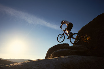 Fototapeta na wymiar Professional sportsman cyclist jumping on trial bicycle on top of rocky mountain. Male rider making acrobatic trick on summer evening, blue sky and sunset on background. Concept of extreme sport