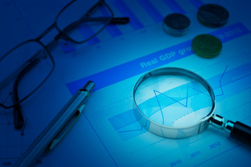 Magnifying glass, pen, coins and glasses on growth financial chart and graph, Business success concept, blue tone
