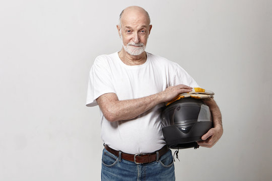 Horizontal image of elderly Caucasian male with thick gray beard posing in studio dressed in white t-shirt and blue jeans holding black motorcycle helmet, looking at camera with confident smile