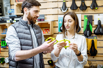 Salesman helping young woman customer to choose paddles standing near the shelves with bicyles parts in the sports shop