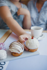 Pink and white marshmallows with curlicues lie next to two cakes on a wooden table against the background of a cup of tea. A hand reaches for the cake.