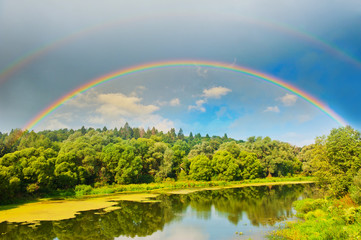 Bright double rainbow in the sky with clouds above the forest and the river