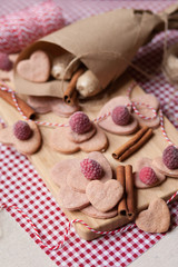 Fototapeta na wymiar Hand made Saint Valentine's day surprise: home made pink sweet cookies in the shape of heart with a frozen raspberries served on a wooden boar and red napkin. Craft paper cornet on a background