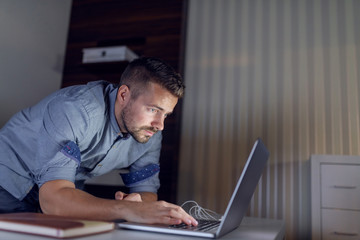 Serious Caucasian freelancer standing in office late at night and typing on keyboard.