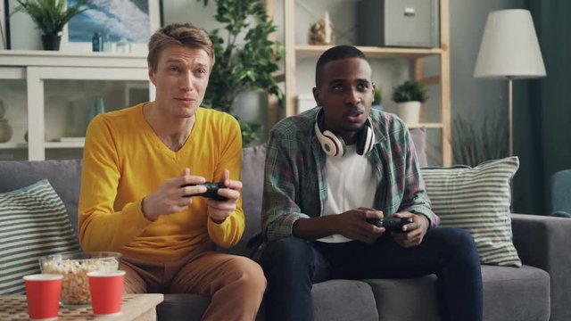 Happy young men are playing video game with console using joysticks st home. Guys are pussing buttons and talking, one is winning, other is losing.
