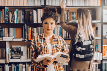 Charming Caucasian brunette with curly hair holding opened book and looking at camera. In background student taking book from shelf. Library interior.
