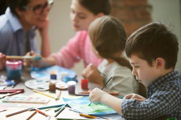 Side view portrait of cute little boy painting pictures in art class with group of children, copy...