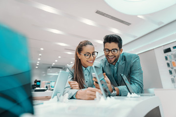 Smiling cute multicultural couple dressed formal and with eyeglasses trying out new smart phone in tech store.