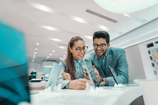 Smiling cute multicultural couple dressed formal and with eyeglasses trying out new smart phone in tech store.
