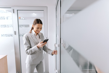 Naklejka na ściany i meble Young Caucasian female CEO dressed in gray formal wear entering the office and using smart phone. For being good leader takes time and experience.