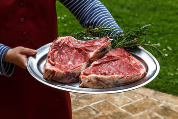 Two big pieces of fresh raw beef meat with rosemary on the plate in the hands on a woman outdoors ready for cooking.