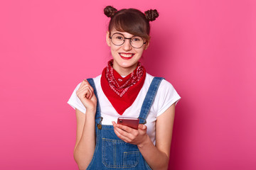 Photo of happy young teenager female with bunches, glasses, bandana on nack, dressed denim overalls...