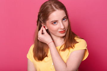 Attractive young girl puts earphones, ready to listen to music, looks derictly at camera, wears bright yellow t shirt, has brown straight hair, spend free time alone. People and entairtaiment concept.