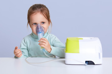 Close up portrait of charming cute small girl uses nebulizer mask for inahlation, has respiratory...