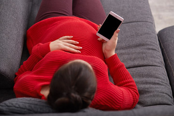 Top view of pregnant female keeps hand on belly, holds mobile phone with blank screen, wears red sweater, with bunch, checking email, lies on comfortable sofa, having rest after going for walk.