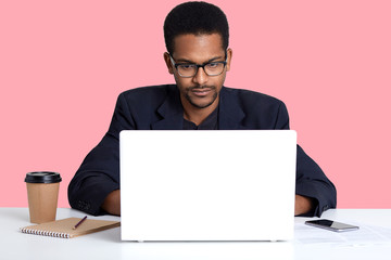 Close up portrait of dark skinned male wears black suit, works online with lap top, African American freelancer sits at white table in front of opened laptop computer, isolated over pink background.