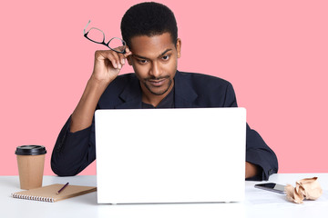Photo of tired man holds glasses in hands near temple, wears black suit, prepares business project, uses modern portable laptop, isolated over pink wall, erans money in office. People concept.