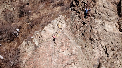 Climbers climb the rock. Drone footage. Side and top view. Rock lesson in the highlands. Gray rocks and dry bushes. Sometimes there is snow. Steep rock and cracks in places.