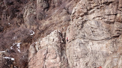 Climbers climb the rock. Drone footage. Side and top view. Rock lesson in the highlands. Gray rocks and dry bushes. Sometimes there is snow. Steep rock and cracks in places.