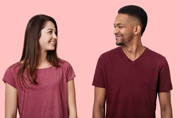 Obraz premium Close up image of happy young couple looking face to face at each other, stands smiling, dressed casual t shirts, enjoys spending time together, pose isolated over pink background in photo studio.