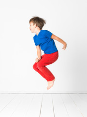 cool, cute boy with blue shirt and red trousers is jumping high in the studio in front of white...