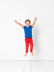 cool, cute boy with blue shirt and red trousers is jumping high in the studio in front of white background and white wooden floor